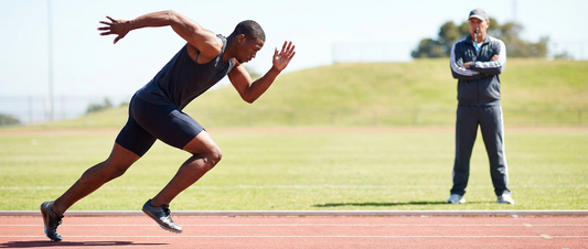 A sprinter running with a coach watching from the background